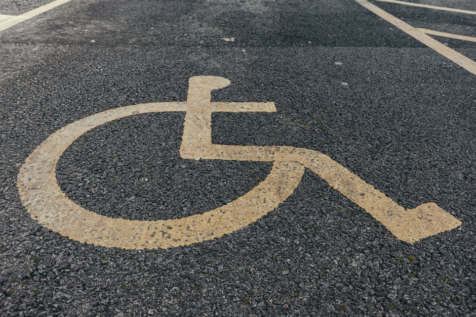 A yellow painted wheelchair accessible parking symbol on a dark asphalt surface, situated in a parking lot near a building. In the background, there are white parking lines and a partially visible vehicle, with some concrete curbing alongside. The parking space is clear of obstructions, and the surface appears slightly textured. This image demonstrates accessible parking facilities, which are relevant to home and business relocations involving parking considerations, as provided by Man with Van Brimsdown, especially when planning access and parking tips for Brimsdown Industrial Estate.