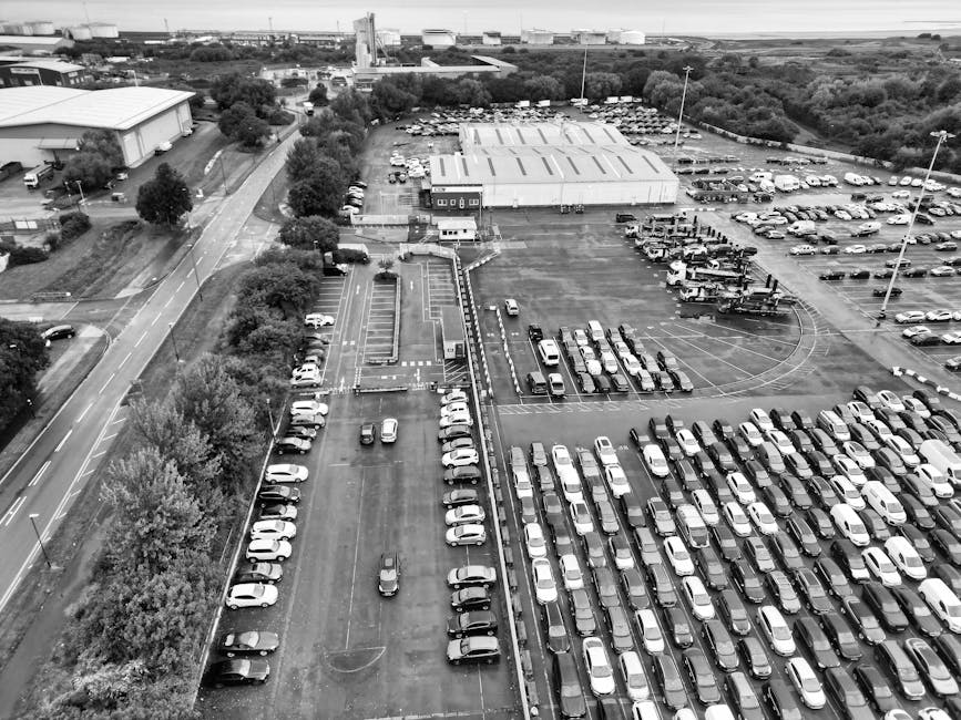 Aerial view of a large parking lot at Brimsdown Industrial Estate showing numerous parked cars arranged in rows on the paved surface, with some vehicles parked close to a warehouse building with a flat roof in the background. To the left, a narrow road runs parallel to the parking area, with a few trees lining its edge. Near the center of the image, a loading zone features a man with van for house removals, visible loading or unloading cardboard boxes, furniture wrapped in plastic or blankets, and other packing materials. The van is parked adjacent to the loading area, with several cardboard boxes and packing supplies nearby. Tall streetlights extend above the parking lot, which is illuminated by natural daylight, and the environment appears organized for efficient vehicle and furniture transport, consistent with professional home relocation services.