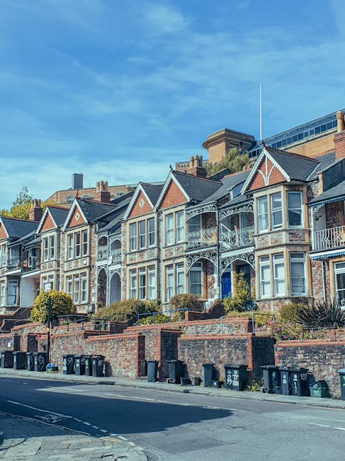 A row of Victorian-style terraced houses with ornate white iron balcony railings, brick facades, and large bay windows, situated behind a brick retaining wall with small landscaped gardens. The houses are positioned on an inclined street with a sidewalk and several black wheelie bins lined along the pavement. The sky above is bright blue with some light clouds, and the scene indicates a residential area typical for home relocation or furniture transport services, as provided by Man with Van Brimsdown. The environment is well-lit, emphasizing the architectural details of the properties and the street scene prepared for moving or packing activities.