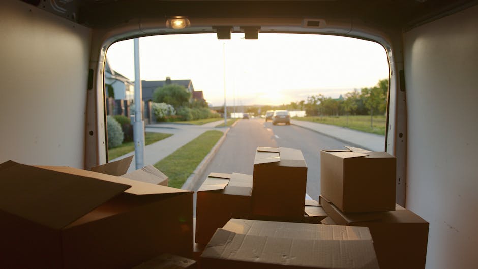 Inside the rear of a moving van, several brown cardboard boxes of various sizes are stacked and arranged on the floor, some partially open, with packing materials visible. The van is parked on a residential street during a clear, sunny late afternoon, with the open back door revealing a view of the pavement, careful roadside gardening, and a few houses in the background. The scene depicts the loading phase of home relocation, with the boxes representing packed belongings ready for transportation. The environment is well-lit with natural daylight, providing a detailed view of the boxes, their arrangement, and the outdoor setting, aligning with professional furniture transport and packing services offered by Man with Van Brimsdown, supporting effective removals and moving logistics.