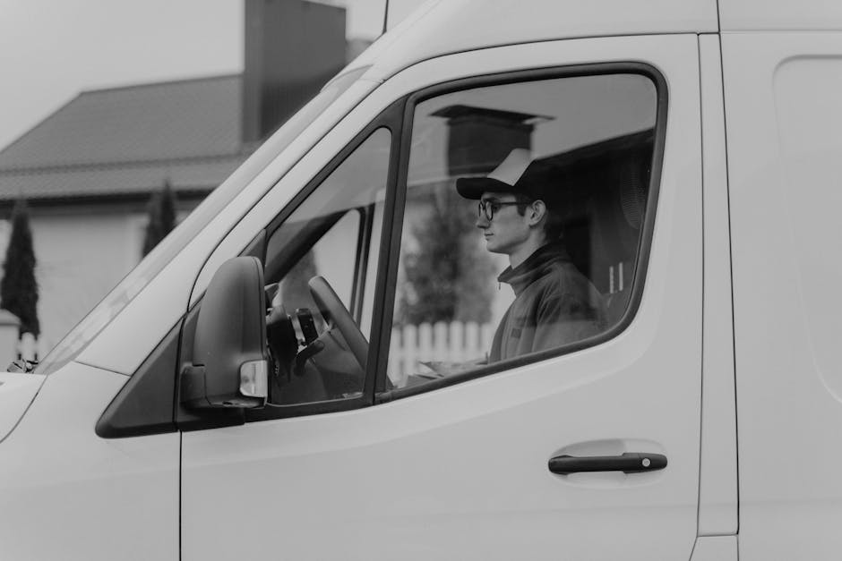 A person wearing a blue beanie hat and a red jacket is in the process of loading several cardboard boxes onto a white, modern commercial van parked in a designated parking area outside a contemporary building with a white facade and large, horizontal windows. The individual is holding one box in their hands while standing on the pavement, with additional boxes placed on the ground nearby, ready for loading. The scene captures a home relocation or furniture transport operation as part of a professional removals service, with the van positioned close to the building's entrance to facilitate efficient packing and moving. The area is well-lit with natural daylight, and the environment appears clean and organized, indicating careful handling of household goods for a house move or interior delivery carried out by [COMPANY_NAME], a specialist in removals and home relocation logistics.