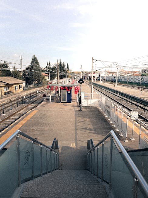 View from the top of a short staircase descending onto a train station platform at Brimsdown, with metal handrails on either side. The platform has a paved surface with designated yellow tactile paving strips along its edge for safety. In the background, there are railway tracks extending into the distance, overhead electric wires supported by tall poles, and a small structure decorated with red and white bunting, which might be used for station staff or ticketing. On the left, a low wooden building is visible, and beyond the station, residential houses and trees can be seen under a partly cloudy sky. The scene is well-lit with natural daylight, illustrating the typical environment for local house removals and furniture transport during a home relocation process by Man with Van Brimsdown, providing access tips for flats and ensuring smooth furniture loading and movement logistics.