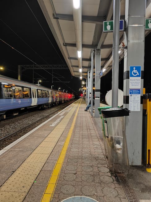 A well-lit outdoor train station platform at night, showing a blue and white passenger train stopped alongside the platform, which is paved with textured tiles and has tactile paving strips near the edge for visually impaired passengers. The platform features a covered waiting area with metal support beams, overhead lighting, and a sign indicating accessible facilities, including a blue wheelchair symbol. A large metal trash bin is positioned close to the sign. In the background, the train extends into the distance under a dark night sky. This scene relates to home relocation logistics, as it illustrates luggage or belongings being prepared for transport, with consideration of loading procedures at stations, as seen in professional removals and packing and moving services provided by Man with Van Brimsdown.