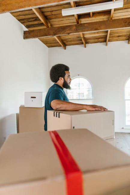 A man with dark curly hair and a beard wearing a navy blue t-shirt is inside a room with white walls and a wooden ceiling, preparing for a home relocation. He is standing behind several large cardboard boxes, some of which are sealed with red packing tape, indicating packing or unpacking processes associated with furniture transport and moving services. Natural light enters through two large arched windows, illuminating the space. The boxes are stacked and placed on a smooth, light-colored floor, with one box positioned directly in front of him and a few stacked behind. The room appears to be an empty or partially furnished interior, suitable for a house removal. The environment suggests an active packing and loading process, with the man possibly about to move the boxes to a vehicle or into another part of the property. The scene aligns with professional moving and packing services offered by Man with Van Brimsdown, supporting efficient furniture transport and logistics within a home relocation context.