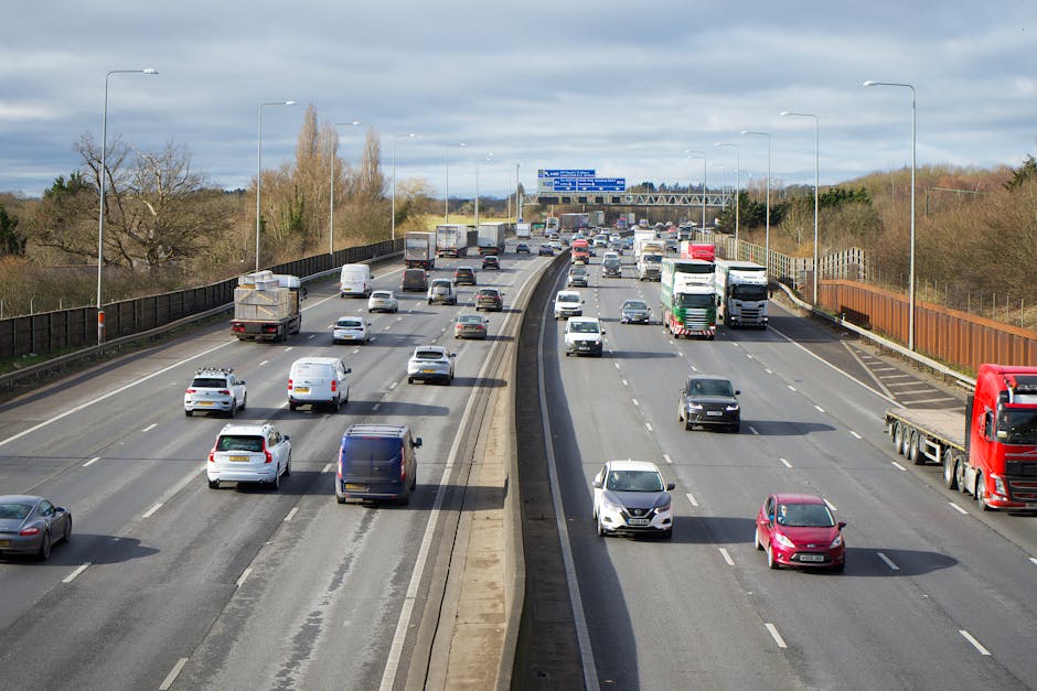 A multi-lane motorway with vehicles including cars, vans, and trucks moving in both directions under a partly cloudy sky. On the left side, a lane barrier separates the carriageway from the opposite traffic, and several additional vehicles can be seen farther along the road. The scene captures the typical flow of traffic associated with home relocation logistics and moving services. Overhead, there are blue directional road signs, and the surrounding scenery features trees with some leaves changing colour, indicating a season transition. The road surface is well-maintained, and the vehicles are in motion, demonstrating the transportation aspect of furniture transport and packing and moving activities. This setting may be relevant to discussions about obtaining permits for moving vans on residential or commercial streets, such as Brimsdown Lane, as managed by Man with Van Brimsdown, an experienced removals company.