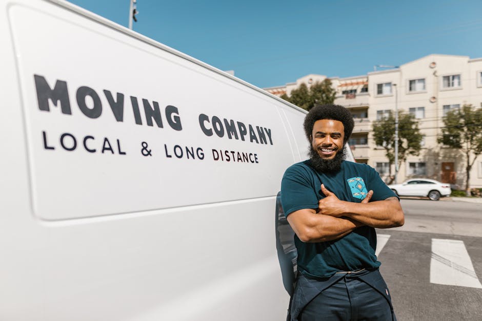 A smiling man with a full beard and an afro hairstyle stands beside a white moving van marked with 'MOVING COMPANY' and 'LOCAL & LONG DISTANCE' on the side. He is wearing a dark blue polo shirt featuring a logo, and his arms are crossed as he leans casually against the van. The background shows a residential street with multi-storey white buildings, parked cars, and trees under a clear blue sky. The van is parked on the pavement, close to the building entrance, suggesting a home relocation or furniture transport activity. Visible elements include cardboard boxes and wrapped furniture inside the open back of the van, indicating the process of packing and loading before a house move. The scene captures a preparation stage typical of professional removals, with the equipment and environment aligned with moving logistics, emphasizing the service provided by Man with Van Brimsdown, specializing in household removals and storage logistics.