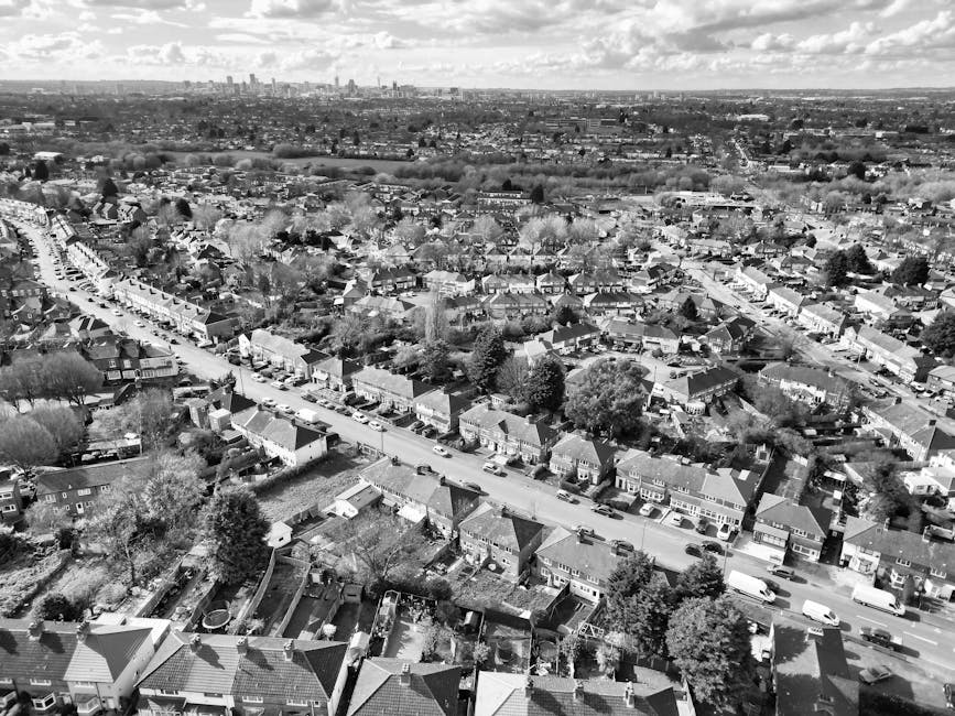 Black and white aerial photograph showing a residential neighbourhood with rows of terraced houses and semi-detached homes along a main street, with parked cars lining the road and garden areas in the foreground. In the background, larger urban buildings and a distant city skyline are visible under a partly cloudy sky. The scene depicts the environment where furniture transport and packing during a home relocation could occur, with the presence of rooftops, trees, and streets suggesting a typical urban suburb. The image provides a broad view suitable for illustrating house removals and moving logistics, such as loading processes or transportation planning, with [COMPANY_NAME] potentially involved in such relocation services within this area, as related to the webpage about emergency same-day removals in EN3: Brimsdown.