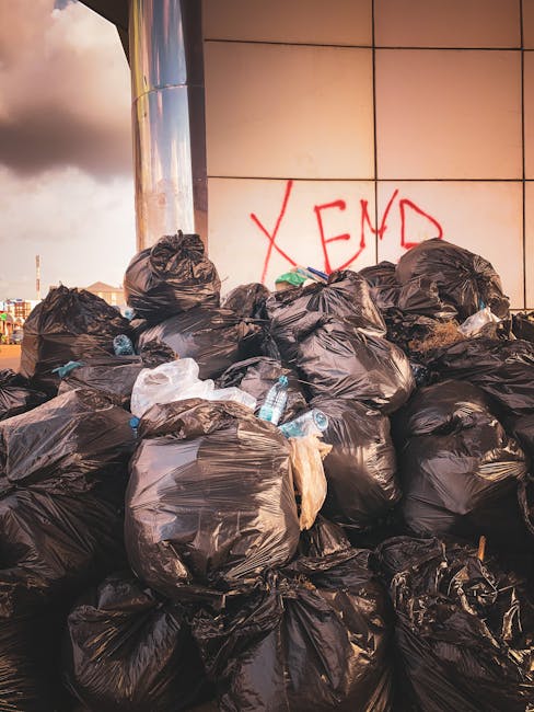 A large pile of black refuse bags filled with waste, some of which contain plastic bottles and paper, is stacked outdoors against a tiled wall with red graffiti reading 'XEND' visible in the background. The bags are placed on pavement, and to the left, a section of a metallic column is visible. The sky above shows clouds with warm sunlight, suggesting late afternoon. This scene illustrates waste disposal near a building, which may relate to the storage or removal process during a house relocation or moving project, as part of managing excess items before a move. Man with Van Brimsdown's services often include assisting clients with removal of unwanted items, ensuring proper disposal as part of home moving or clearance tasks.