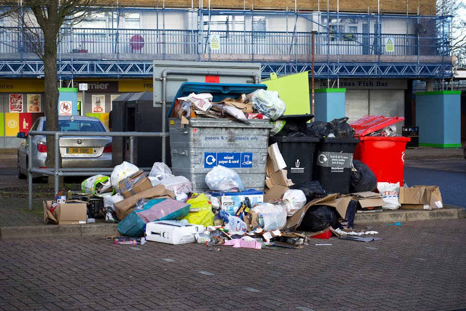 A collection of overflowing public waste and recycling bins situated on a paved sidewalk in an urban area, with various items including cardboard boxes, plastic bags, paper packaging, and black trash bags scattered around the bins and on the ground. The bins are of different colours and sizes, with some lids open and trash spilling out. Behind the bins, there is a metal railing, and a grey car parked nearby. In the background, there is a building with a blue scaffolding structure and retail shopfronts, indicating a commercial or shopping area. The scene illustrates improper waste disposal, which local authorities such as Man with Van Brimsdown may assist with through house and business removal services that include responsible waste management and relocation of belongings, supporting proper disposal practices during home relocations or removals.