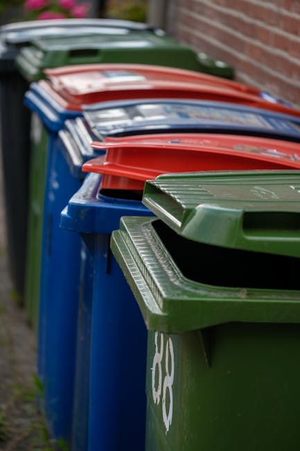 A large pile of black refuse bags filled with waste, some of which contain plastic bottles and paper, is stacked outdoors against a tiled wall with red graffiti reading 'XEND' visible in the background. The bags are placed on pavement, and to the left, a section of a metallic column is visible. The sky above shows clouds with warm sunlight, suggesting late afternoon. This scene illustrates waste disposal near a building, which may relate to the storage or removal process during a house relocation or moving project, as part of managing excess items before a move. Man with Van Brimsdown's services often include assisting clients with removal of unwanted items, ensuring proper disposal as part of home moving or clearance tasks.