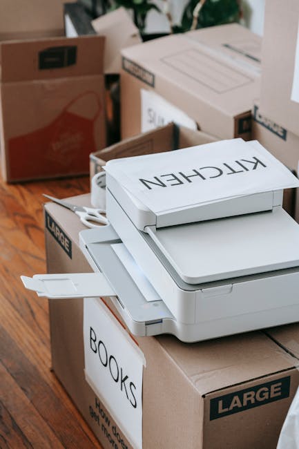 A young man with dark hair, wearing a white t-shirt and blue jeans, is sitting on the floor inside a property surrounded by numerous cardboard boxes of various sizes, some sealed with packing tape and