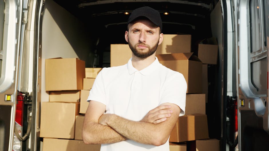 A young man with a beard, wearing a red cap backwards, a checkered shirt, beige and brown jacket, and striped gloves, sitting on the open tailgate of a white moving van. Behind him inside the van are