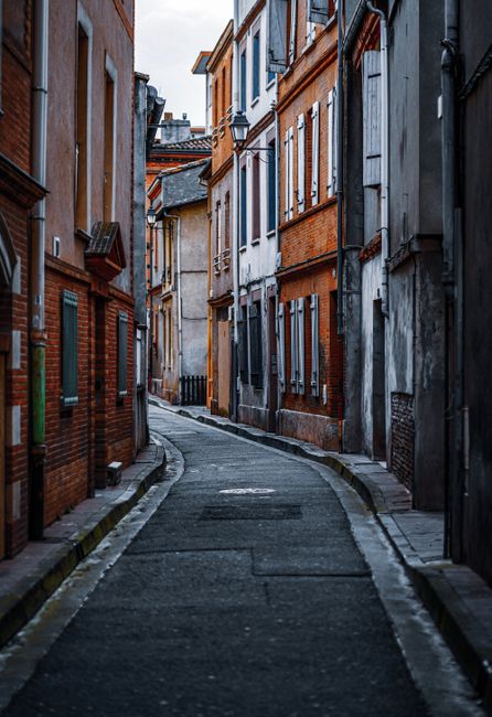A narrow, winding street lined with multi-storey brick and stone terraced houses featuring various windows and doorways, some with shutters or small awnings, in an urban residential area. The street is paved with asphalt and has a slight curve, with a mix of red, brown, and gray building facades. There are no people or vehicles visible. This setting illustrates a typical home relocation environment, suitable for describing the challenges of narrow street moving solutions, where careful loading and transport planning is essential for house removals. Occasionally, Man with Van Brimsdown services are involved in managing the logistics of furniture transport and packing through tight urban streets, ensuring efficient and safe moving processes in confined spaces.
