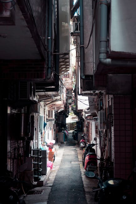 A narrow residential alleyway with closely packed multi-storey buildings on both sides, featuring external staircases, air conditioning units, and exposed pipes on the exterior walls. The alley has a paving surface with various items such as cardboard boxes, plastic containers, and small furniture pieces placed along the sides, suggesting a moving or packing process. Motorcycles and bicycles are parked against the walls, and a few people are visible in the background, possibly involved in a home relocation. Overhead, there are electrical wires extending between the buildings. The scene is well-lit, indicating daylight, with a focus on the confined space suitable for small-scale removals or furniture transport, aligning with the services offered by Man with Van Brimsdown for tight-street removals in EN3, focusing on narrow terrace solutions and moving logistics.